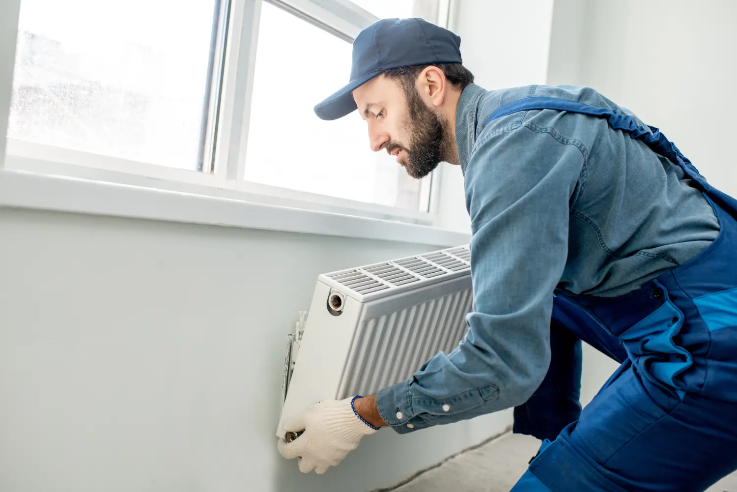 Handwerker montiert Warmwasserheizkörper in der Nähe des Fensters im weiß renovierten Wohnzimmer